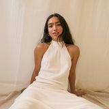 Woman wearing a sun dangle earrings and a white halter neck dress sitting against a beige curtain.
