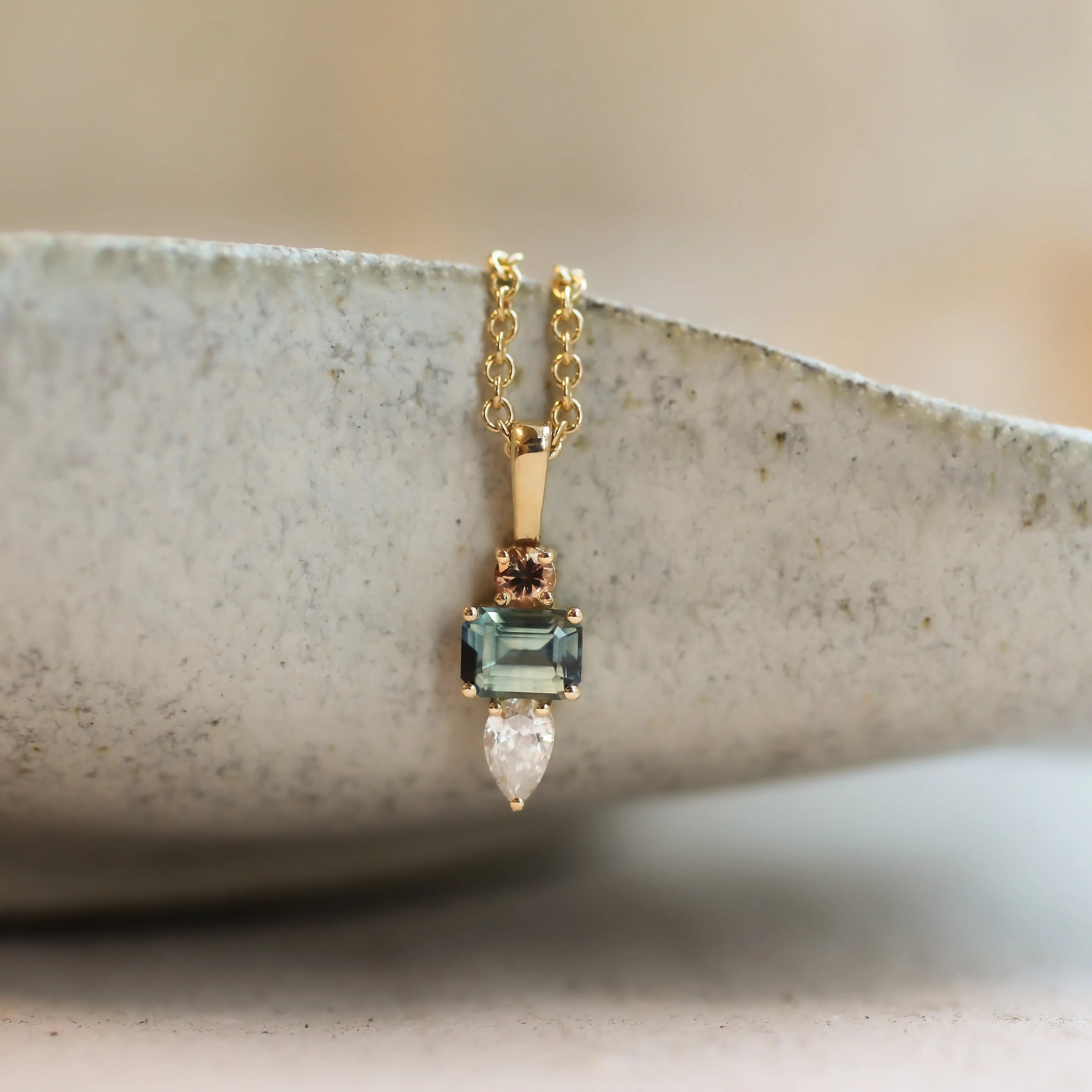 A delicate gold pendant necklace with a light blue-green rectangular gemstone, a small rose-gold accent, and a clear teardrop-shaped diamond, resting on the rim of a textured ceramic bowl.