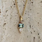 A gold pendant necklace featuring a rectangular light blue gemstone, a marquise-cut clear stone, and a small rose gold accent, displayed against a textured beige stone background.