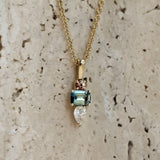 A gold pendant necklace featuring a rectangular light blue gemstone, a marquise-cut clear stone, and a small rose gold accent, displayed against a textured beige stone background.