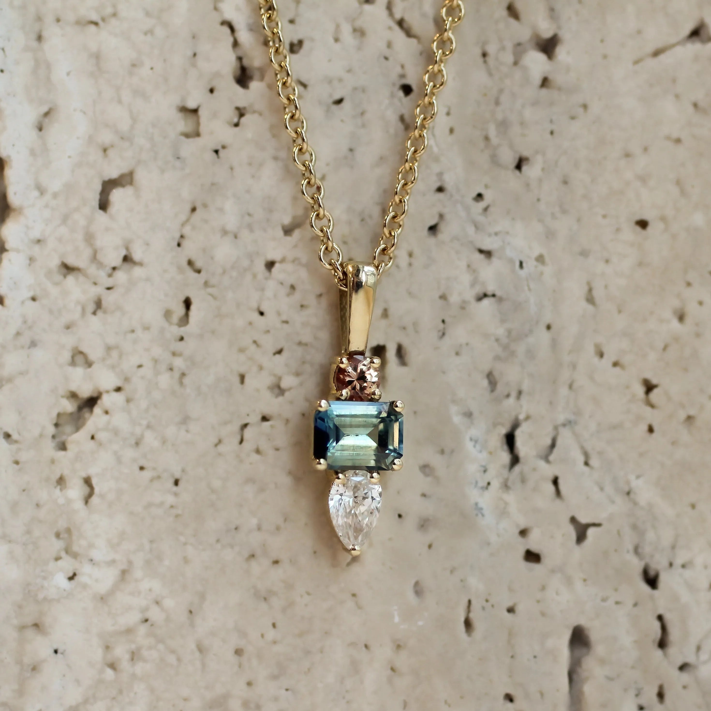A gold pendant necklace featuring a rectangular light blue gemstone, a marquise-cut clear stone, and a small rose gold accent, displayed against a textured beige stone background.
