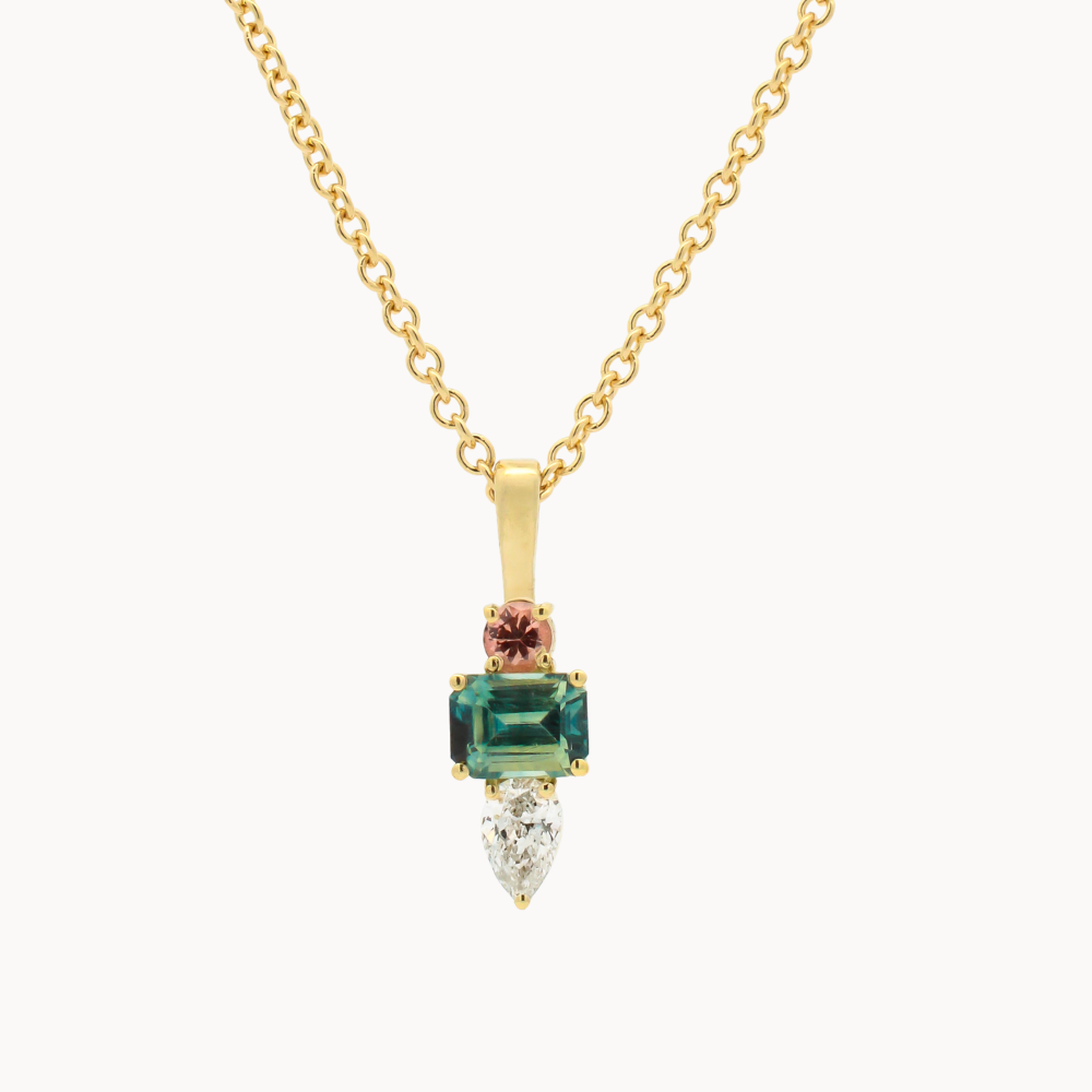 A gold pendant necklace featuring a blue/green emerald-cut sapphire, a pear diamond, and a small round pink gemstone on a delicate gold chain, set against a plain light background.