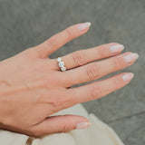 A close-up of a hand wearing a gold ring with three clear round-cut gemstones, set against a soft gray textured background.