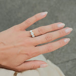 A close-up of a hand wearing a gold ring with three clear round-cut gemstones, set against a soft gray textured background.