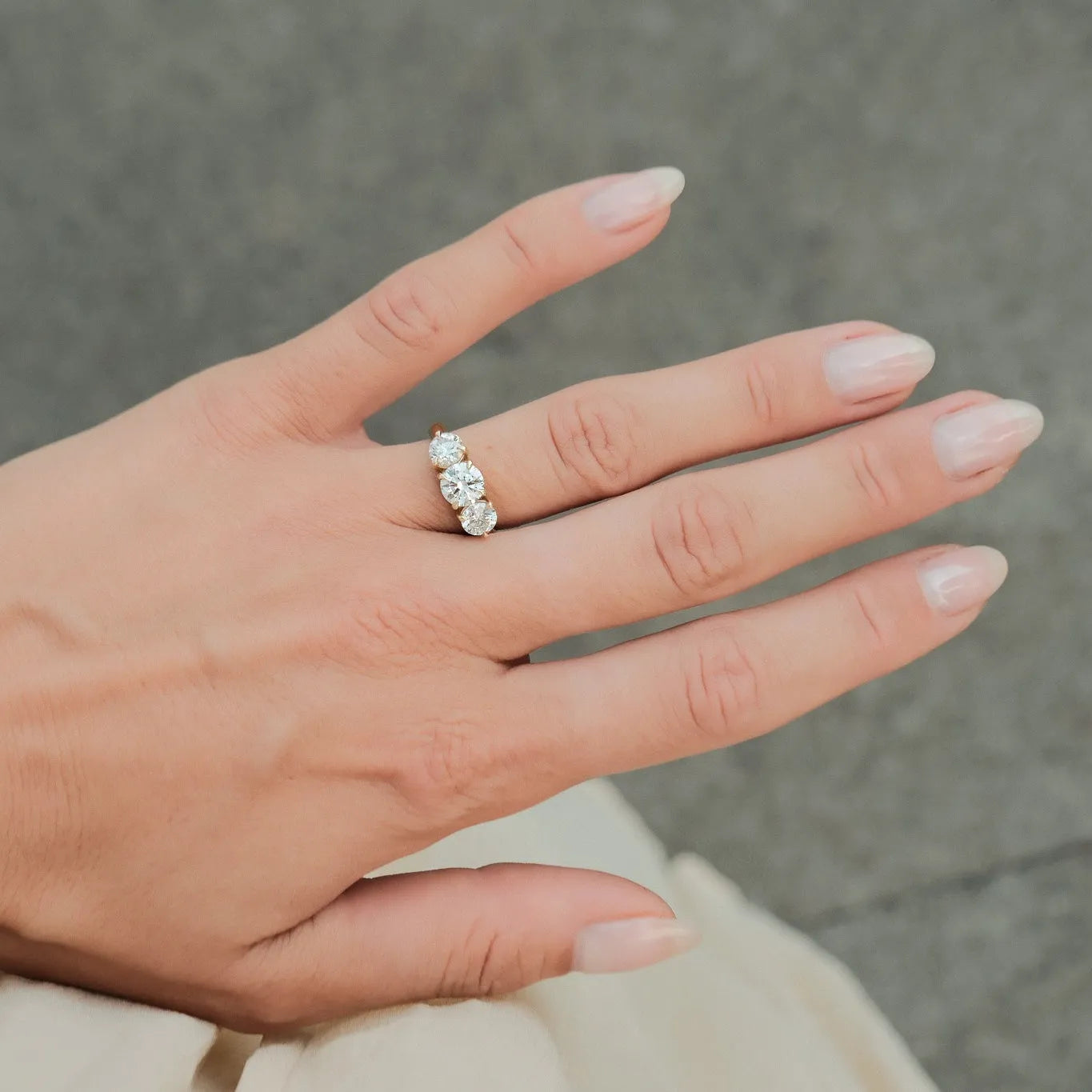 A close-up of a hand wearing a gold ring with three clear round-cut gemstones, set against a soft gray textured background.