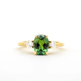 A yellow gold ring featuring a central oval-cut green gemstone flanked by two small round clear stones, set against a plain white background.