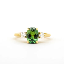 A yellow gold ring featuring a central oval-cut green gemstone flanked by two small round clear stones, set against a plain white background.