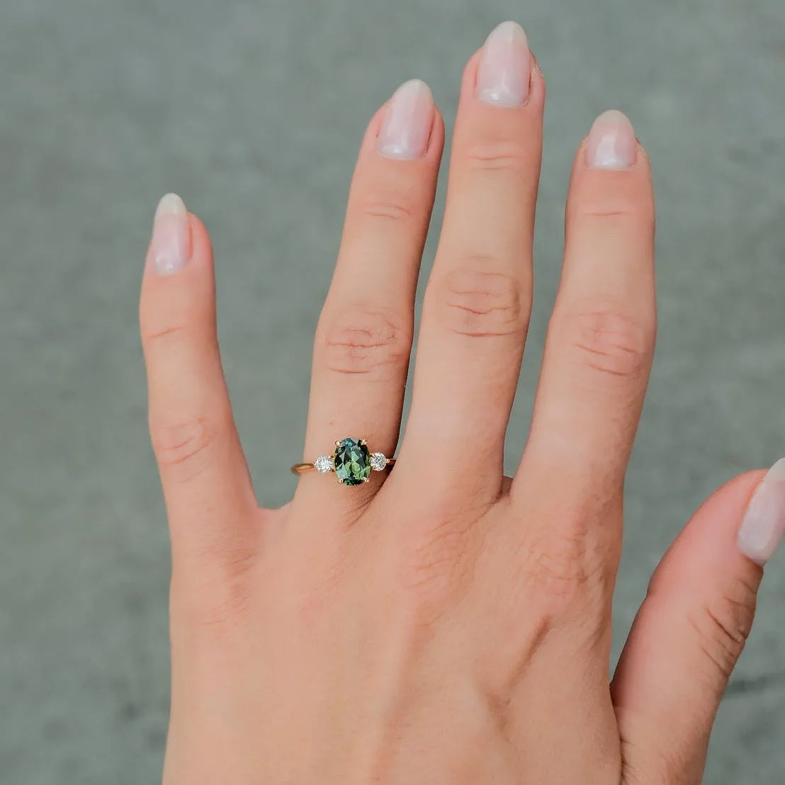 A close-up of a person's hand wearing a gold ring with a green gemstone, set against a gray paved background.
