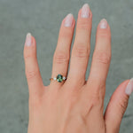 A close-up of a person's hand wearing a gold ring with a green gemstone, set against a gray paved background.