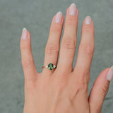 A close-up of a person's hand wearing a gold ring with a green gemstone, set against a gray paved background.
