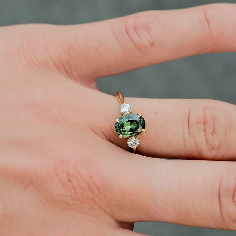 A close-up of a hand wearing a ring with a green oval center stone and small clear accent stones on a gold-toned band, set against a blurred gray background.
