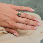 A close-up of a person's hand wearing a gold ring with a green and white gemstone, resting on draped beige fabric against a gray textured background.