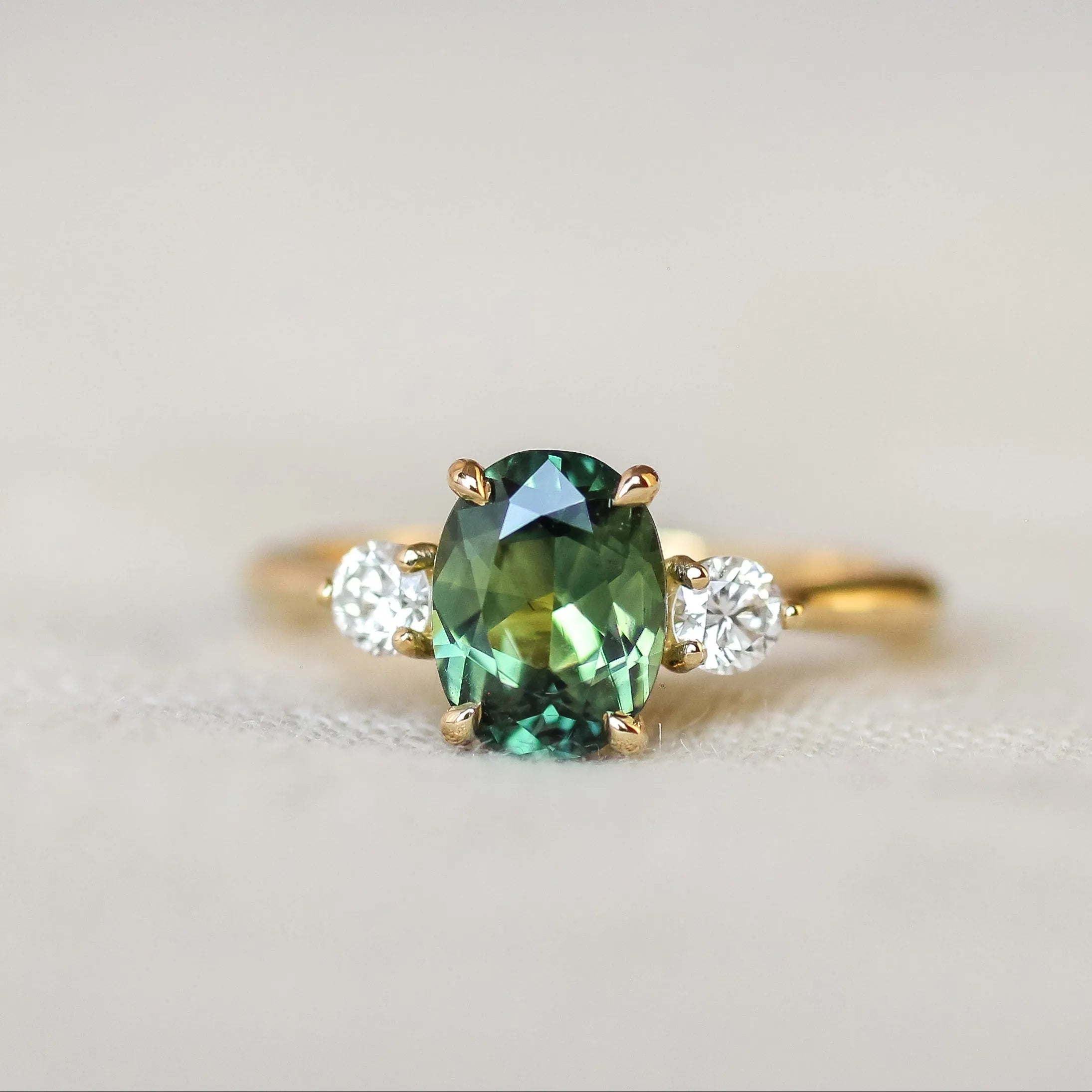 A gold ring featuring a central oval-cut green gemstone flanked by two small round clear stones, photographed on a soft light beige surface.