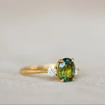 A gold ring featuring an oval green gemstone in the center and a small clear accent stone on each side, shown on a soft beige surface.
