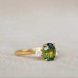 A gold ring featuring an oval green gemstone in the center and a small clear accent stone on each side, shown on a soft beige surface.