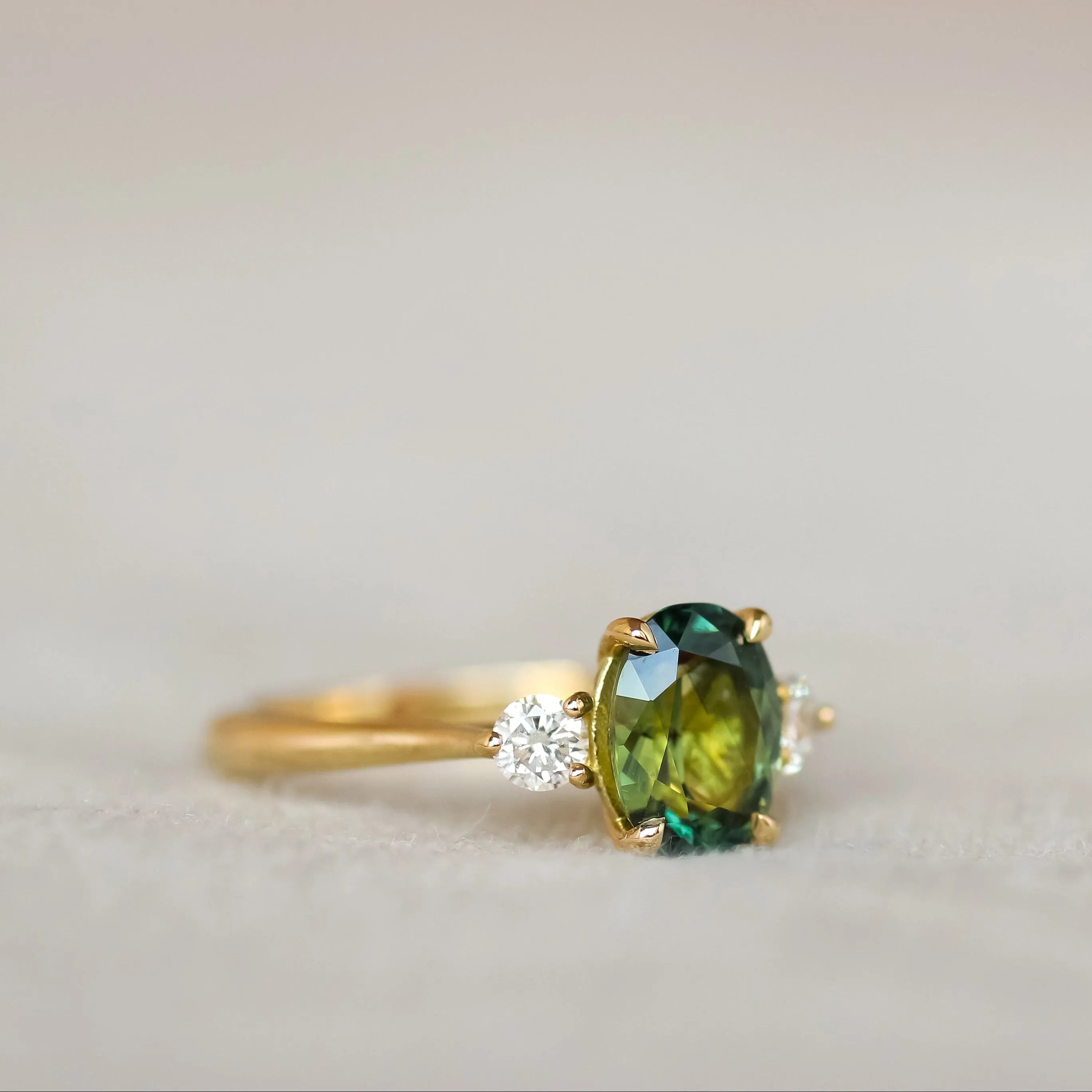 A gold ring featuring an oval green gemstone in the center and a small clear accent stone on each side, shown on a soft beige surface.