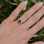 A close-up of a hand wearing a ring with a round green center stone and small clear side stones, photographed outdoors against a blurred green background.