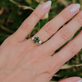 A close-up of a hand wearing a ring with a round green center stone and small clear side stones, photographed outdoors against a blurred green background.