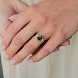 A close-up of a person's hand wearing a gold ring with a round green gemstone and small clear accent stones, set against a soft cream-colored background.