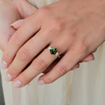 A close-up of a person's hand wearing a gold ring with a round green gemstone and small clear accent stones, set against a soft cream-colored background.
