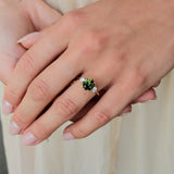 A close-up of a person's hand wearing a gold ring with a round green gemstone and small clear accent stones, set against a soft cream-colored background.