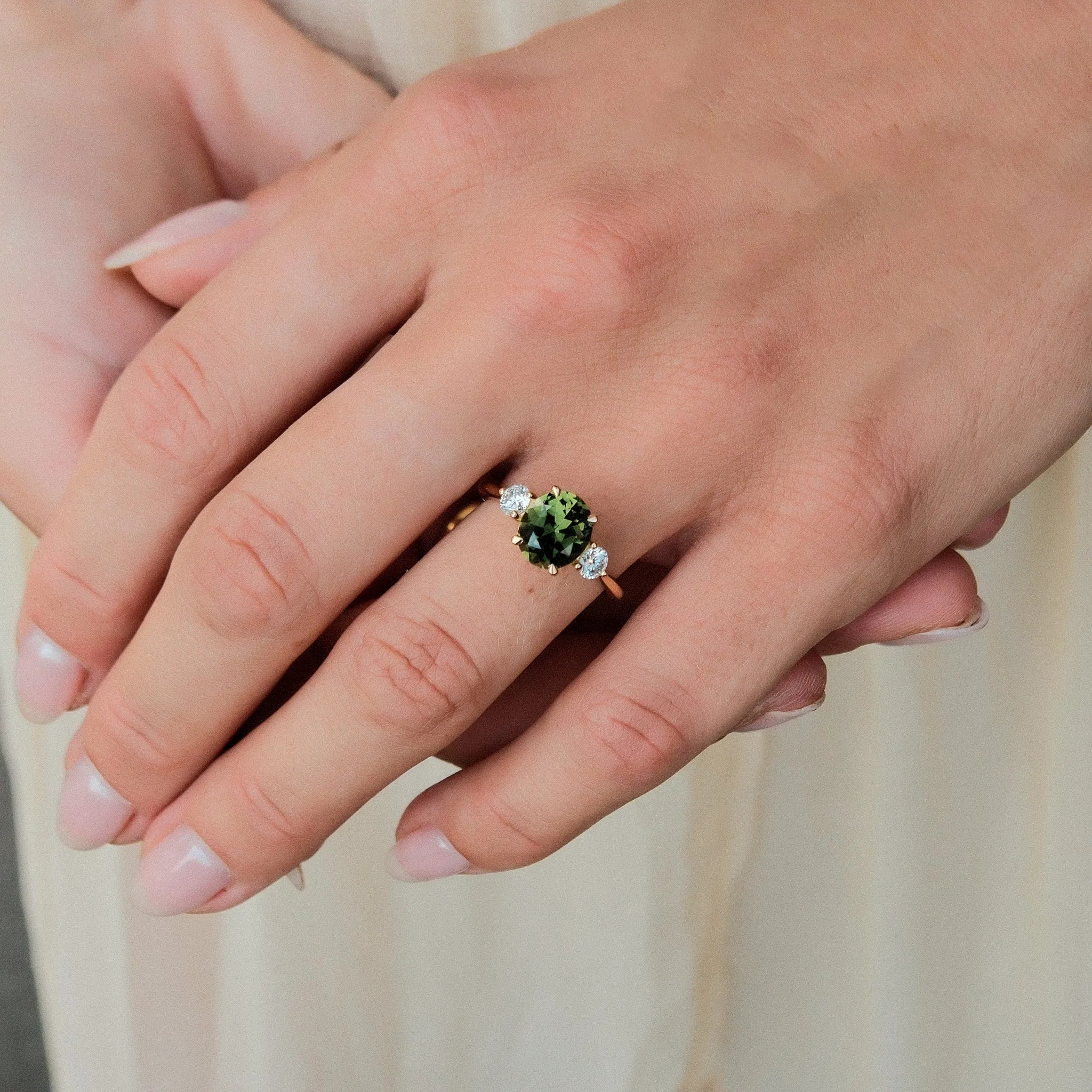 A close-up of a person's hand wearing a gold ring with a round green gemstone and small clear accent stones, set against a soft cream-colored background.