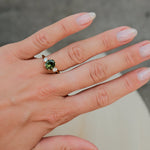 A close-up of a hand wearing a silver ring with a green gemstone, set against a soft gray background.