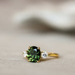 A gold ring featuring a round green gemstone in the center, flanked by two small clear accent stones, resting on a soft beige surface with a blurred white background.