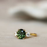 A gold ring featuring a round green gemstone in the center, flanked by two small clear accent stones, resting on a soft beige surface with a blurred white background.