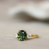 A gold ring featuring a round green gemstone in the center, flanked by two small clear accent stones, resting on a soft beige surface with a blurred white background.