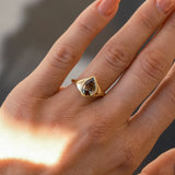 A gold ring with a teardrop-shaped brown gemstone displayed on a person's finger, captured in warm sunlight against a blurred gray background.