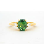 yellow gold engagement ring with a bright green oval sapphire flanked by round diamonds, against a plain white background
