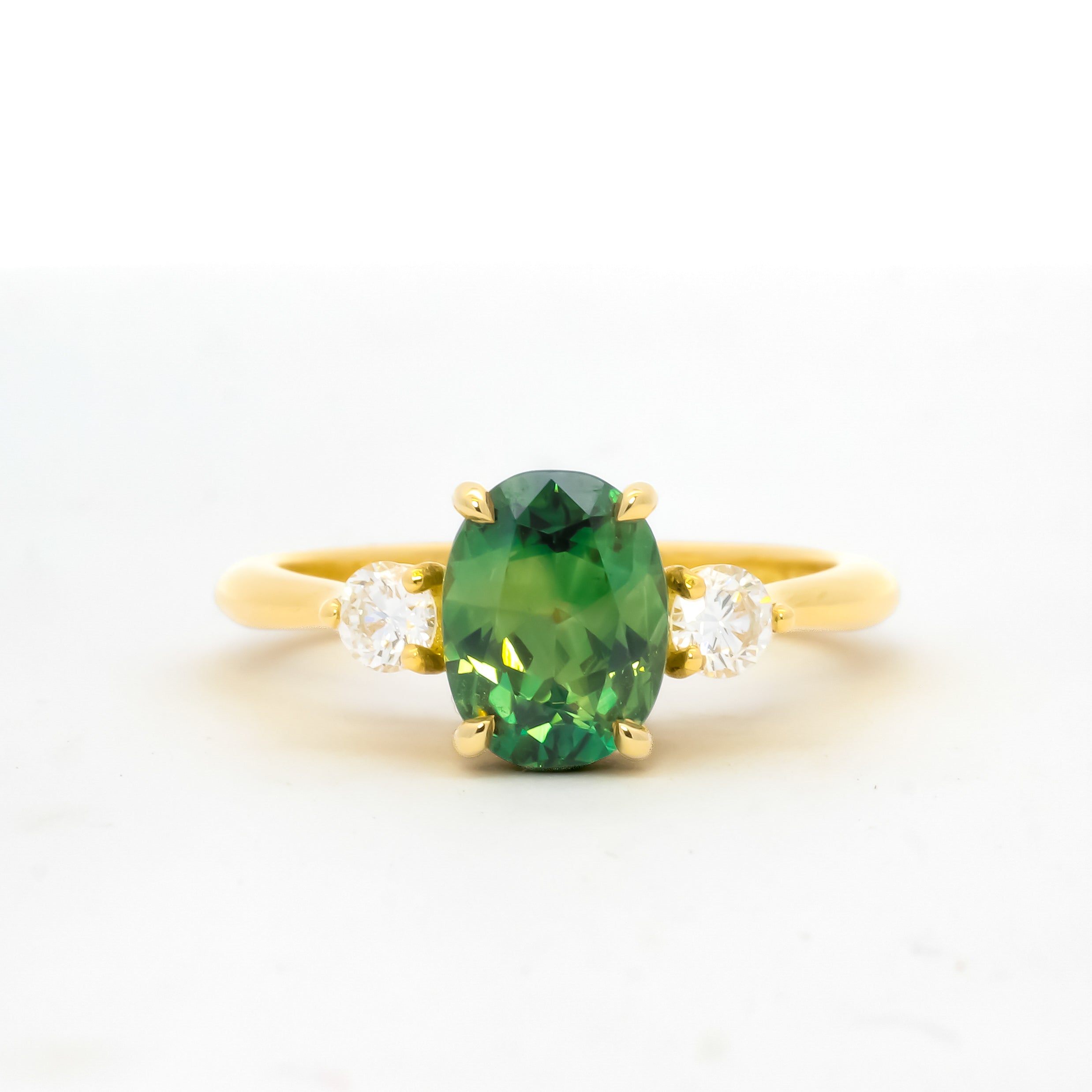 yellow gold engagement ring with a bright green oval sapphire flanked by round diamonds, against a plain white background