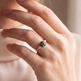 A close-up lifestyle shot of a person wearing a gold ring with a green gemstone and kite-shaped diamonds, dressed in a cream-colored turtleneck sweater.