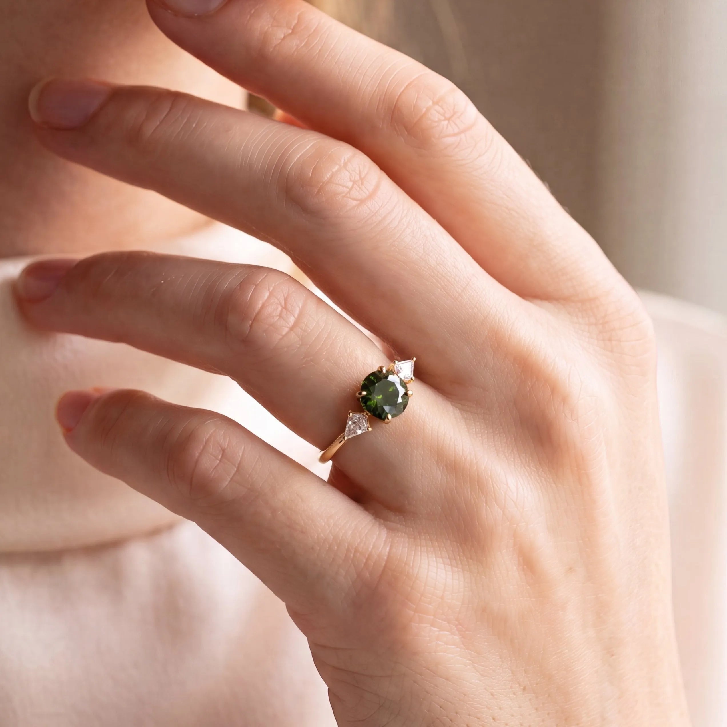 A close-up lifestyle shot of a person wearing a gold ring with a green gemstone and kite-shaped diamonds, dressed in a cream-colored turtleneck sweater.