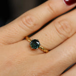 Gold twisted engagement ring with a green gemstone on a hand against a dark background