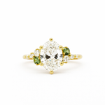 cluster engagement ring with large oval diamonds, smaller round diamonds and two green small sapphires in gold on a white background