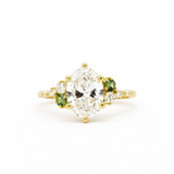 cluster engagement ring with large oval diamonds, smaller round diamonds and two green small sapphires in gold on a white background