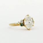 Gold engagement ring with a large diamond and smaller green gemstones on a white background