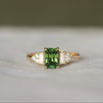 Gold engagement ring with a green rectangular sapphire gemstone and diamond cluster on a white fabric background