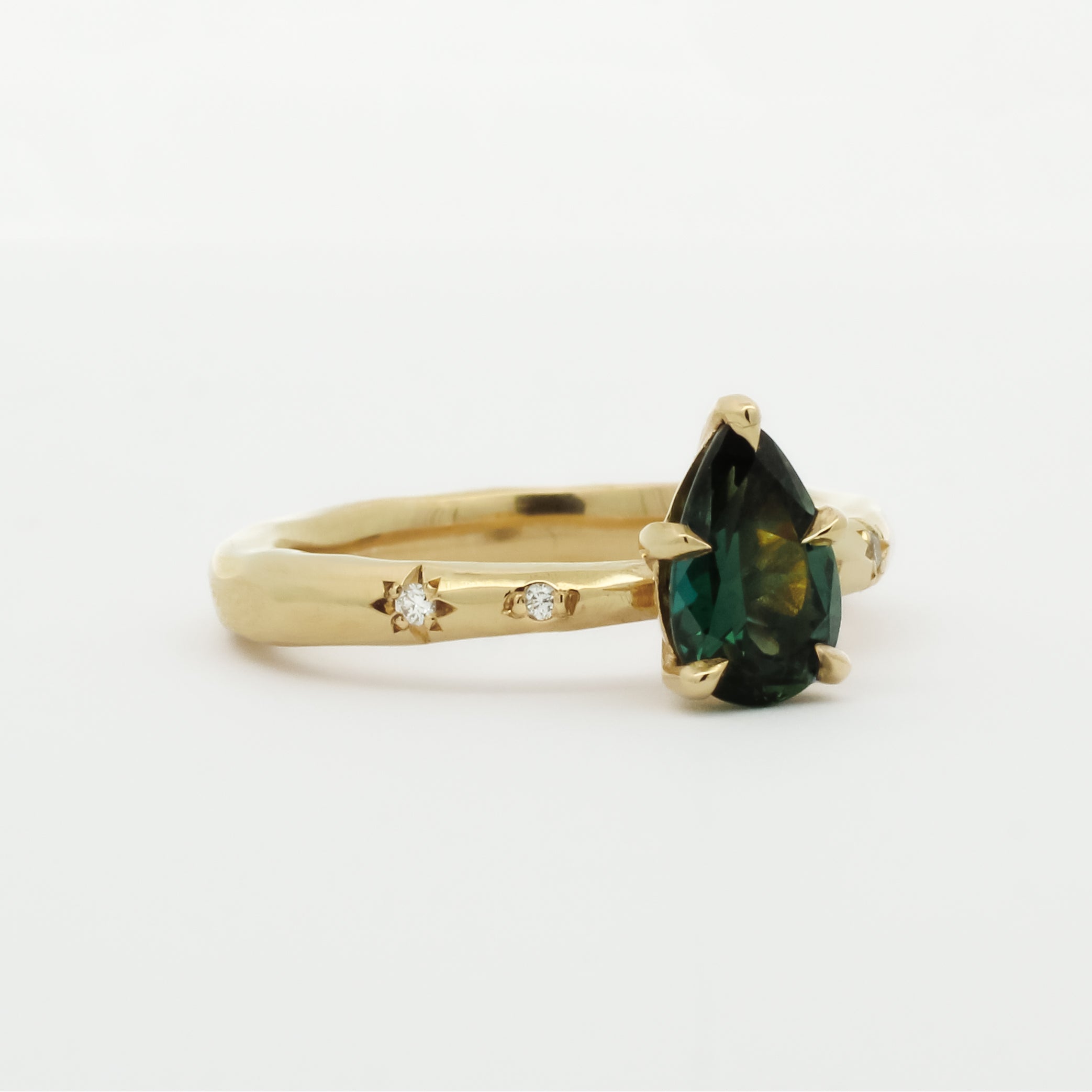 side view of gold engagement ring with organic band, star-set diamonds and a green pear gemstone on a white background