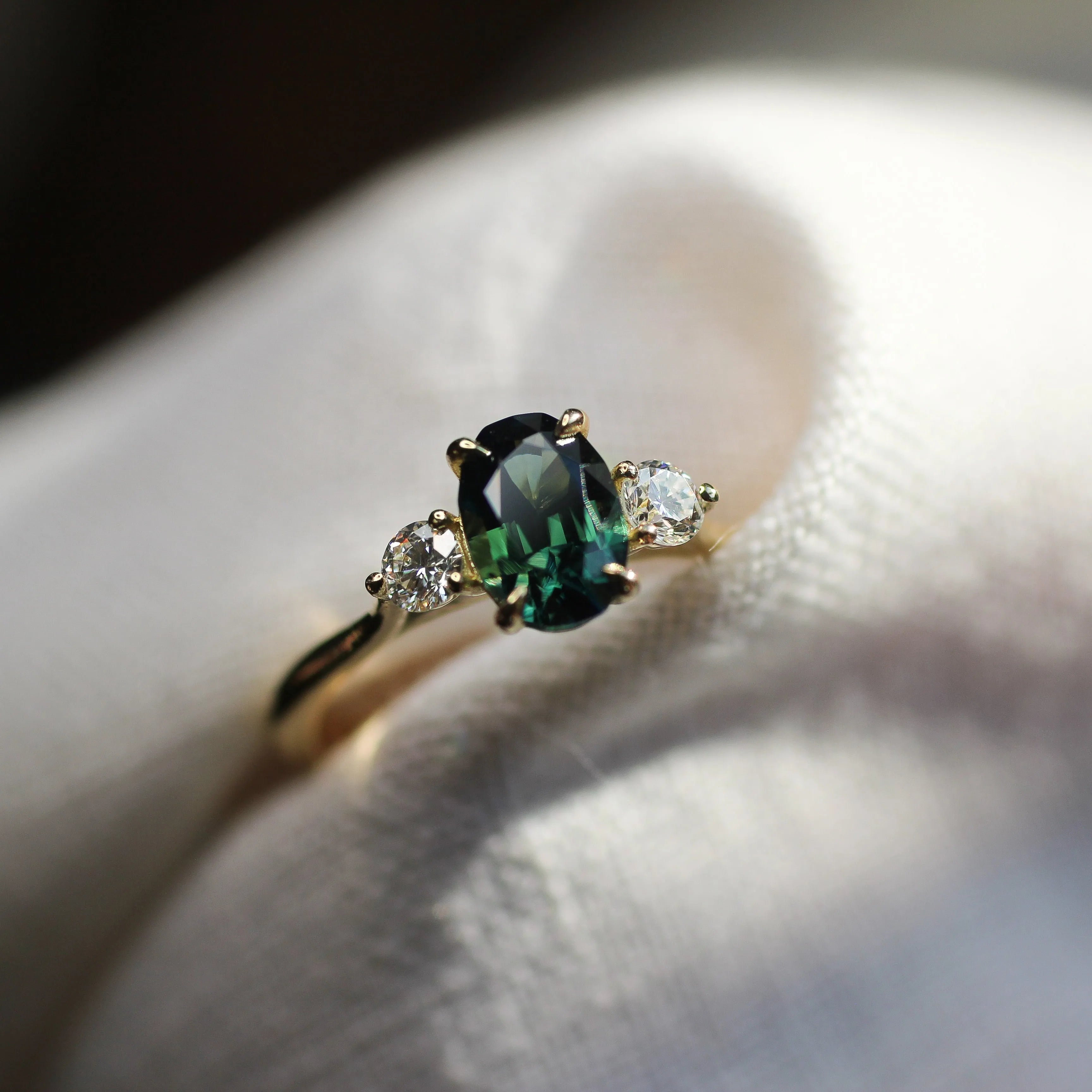 trilogy engagement ring with peacock blue green oval sapphire in blurred neutral fabric