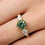 round parti green blue sapphire engagement ring with round diamonds close up being worn on a finger