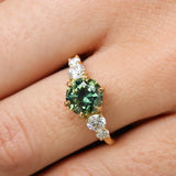 round parti green blue sapphire engagement ring with round diamonds close up being worn on a finger
