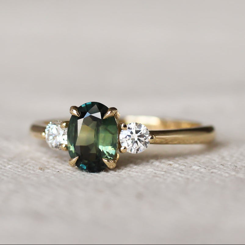 Gold trilogy engagement ring with a oval green gemstone and round diamonds on a textured surface
