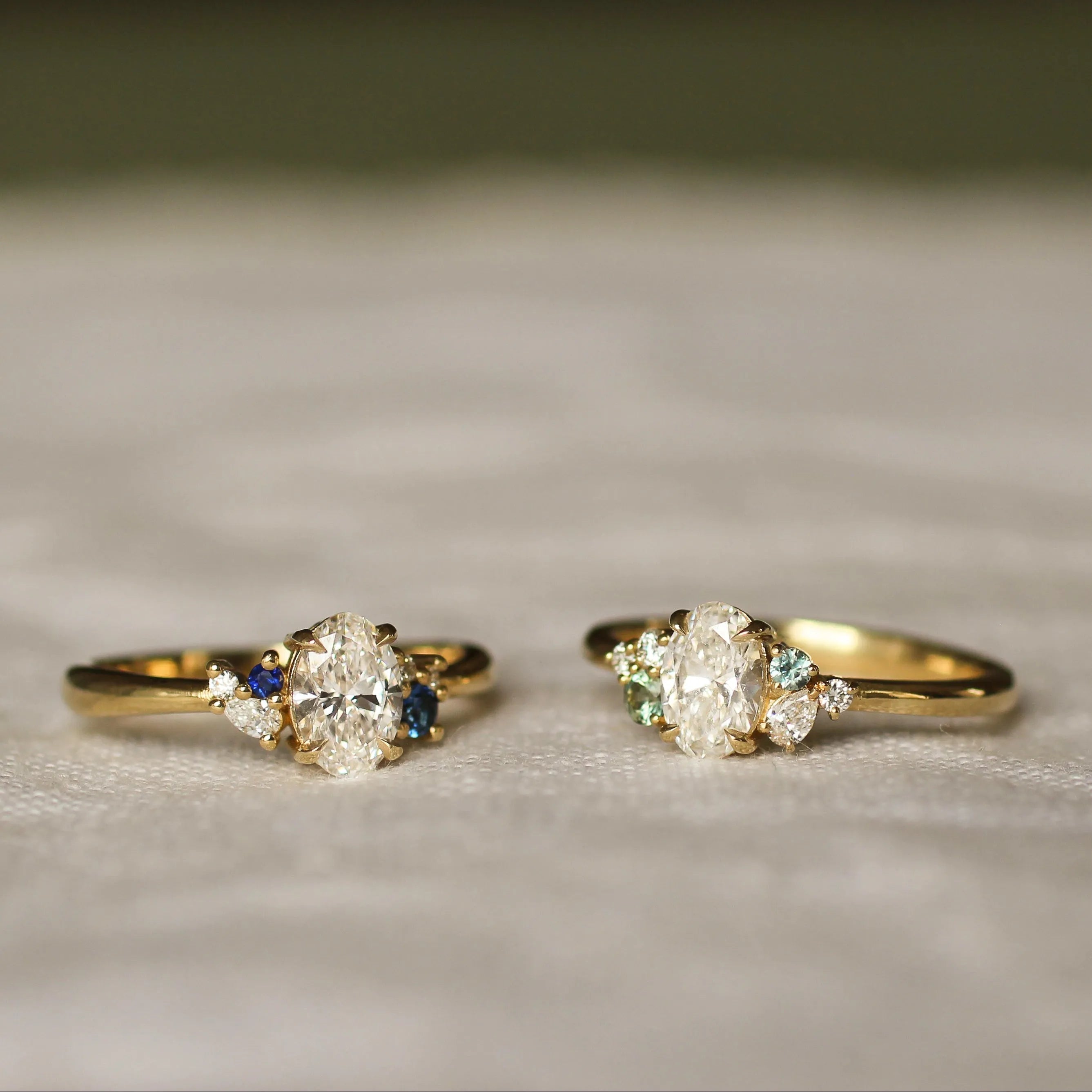 Two gold rings with diamond and blue and green sapphire stones on a textured surface