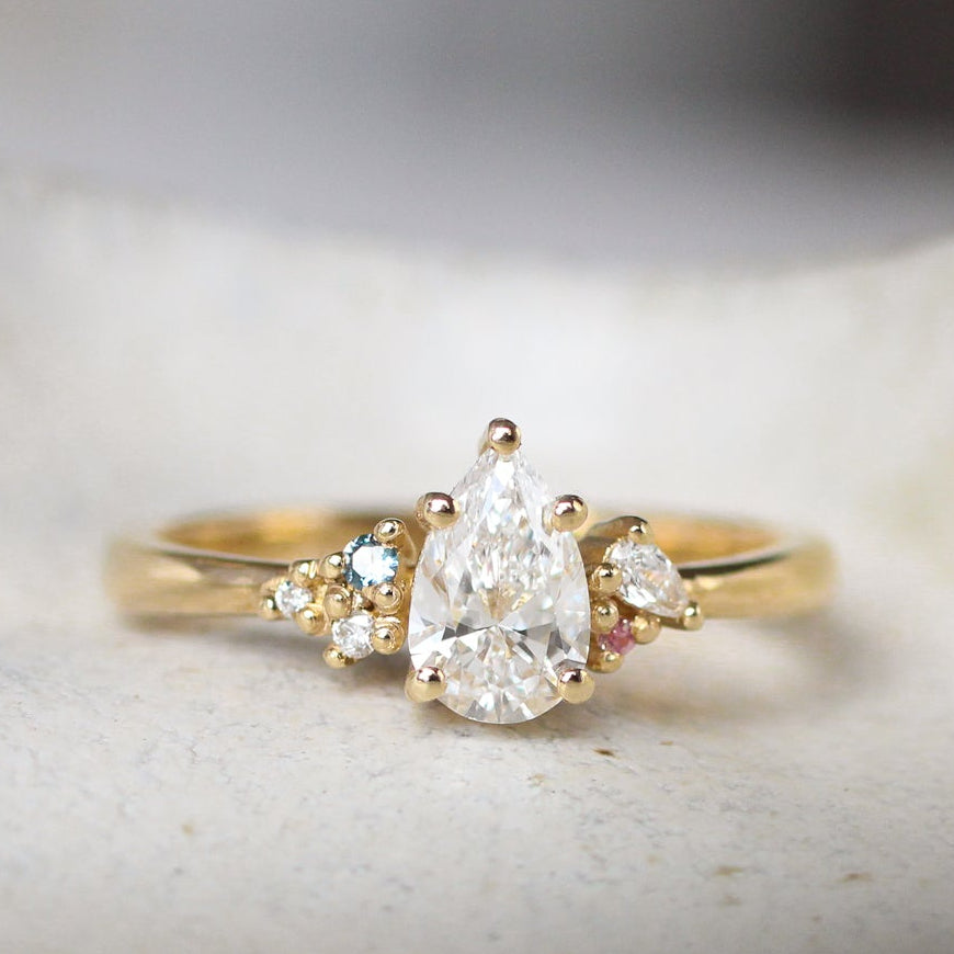 Gold ring with a pear-shaped diamond and small blue and pink sapphires on a textured surface