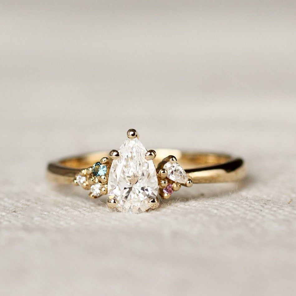 Gold ring with a pear diamond and small blue and pink sapphires on a light background