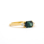 side view of east-west trilogy engagement ring with emerald-cut rectangular parti-sapphire and round diamonds on a white background