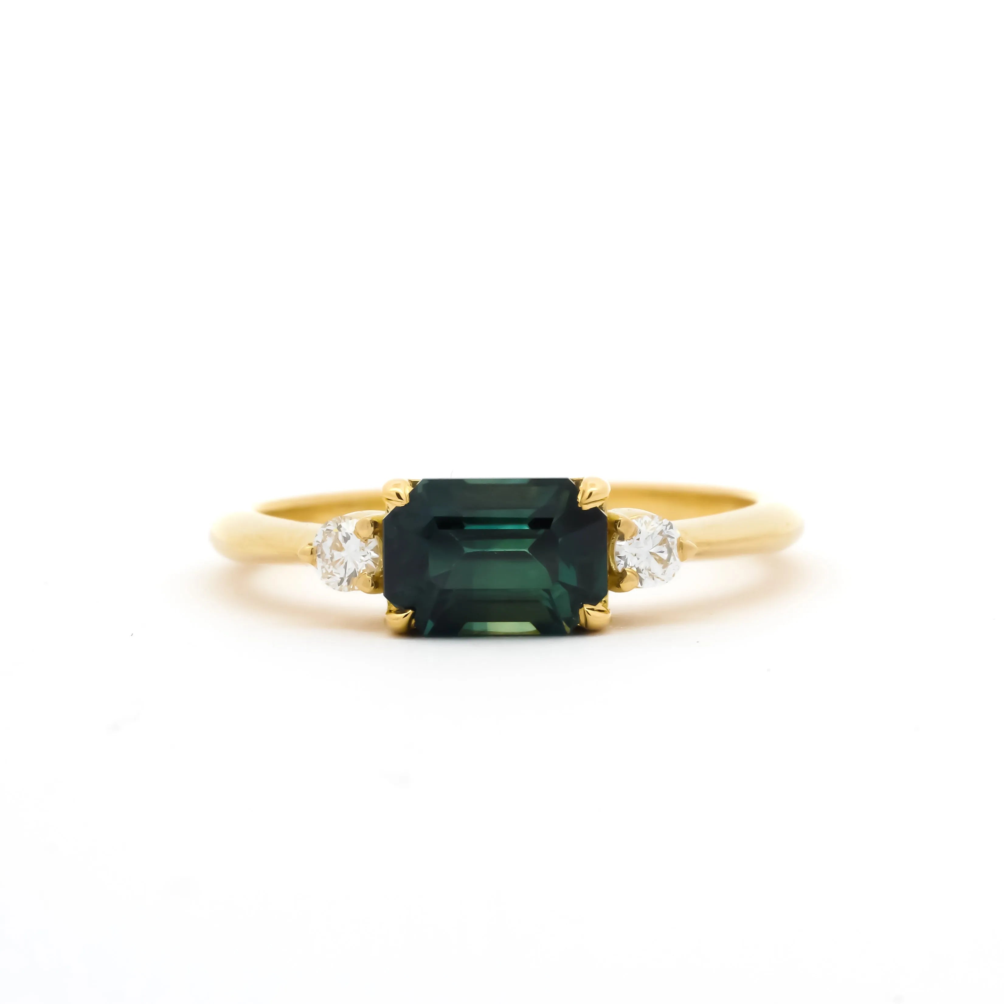 Gold engagement ring with a east-west emerald-cut blue green parti sapphire and diamonds on a white background